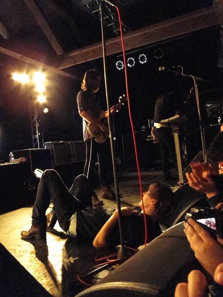 Thomas Mars lying on a stage floor, the band playing above him under bright lights