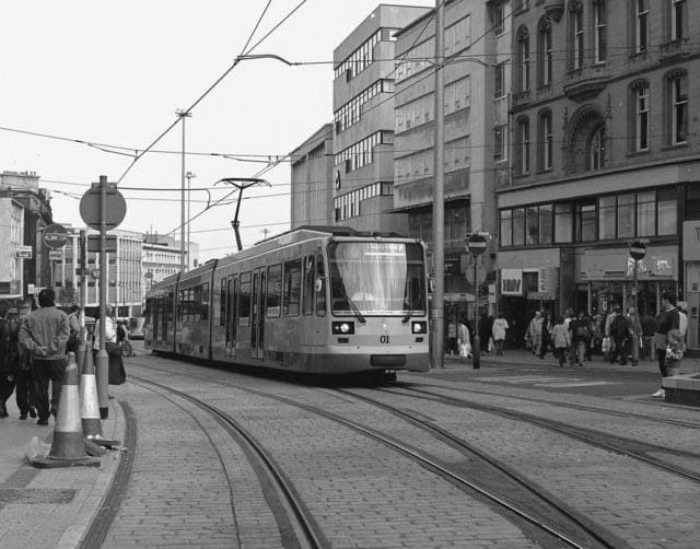 Black-and-white photograph of a Supertram climbing a city-centre street in Sheffield in 1995, pedestrians on the pavement beside cones