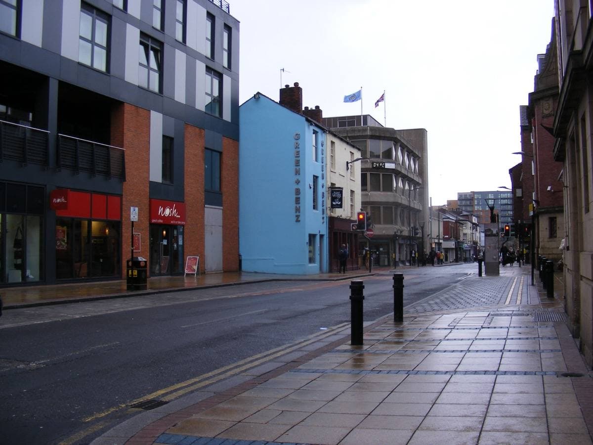 Division Street, Sheffield, on a wet day: shopfronts, a tall blue building and new flats along a broad pavement