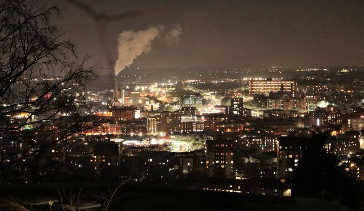 Sheffield at night from a hillside: a bowl of orange street lights and office windows, a white plume rising from a chimney, bare branches in the foreground