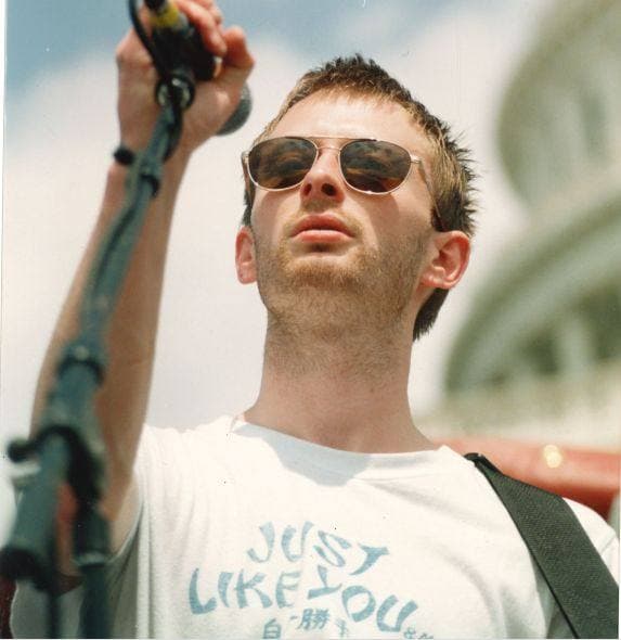 Thom Yorke in sunglasses and a white T-shirt at a microphone, a domed building behind