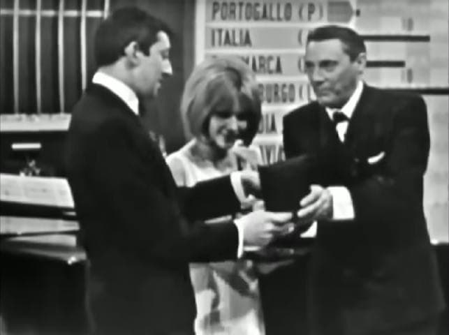 A television frame of Serge Gainsbourg in a dark suit facing France Gall and Mario Del Monaco on a stage
