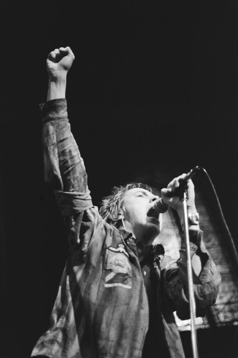 Johnny Rotten singing with one fist raised, in a checked shirt, black and white