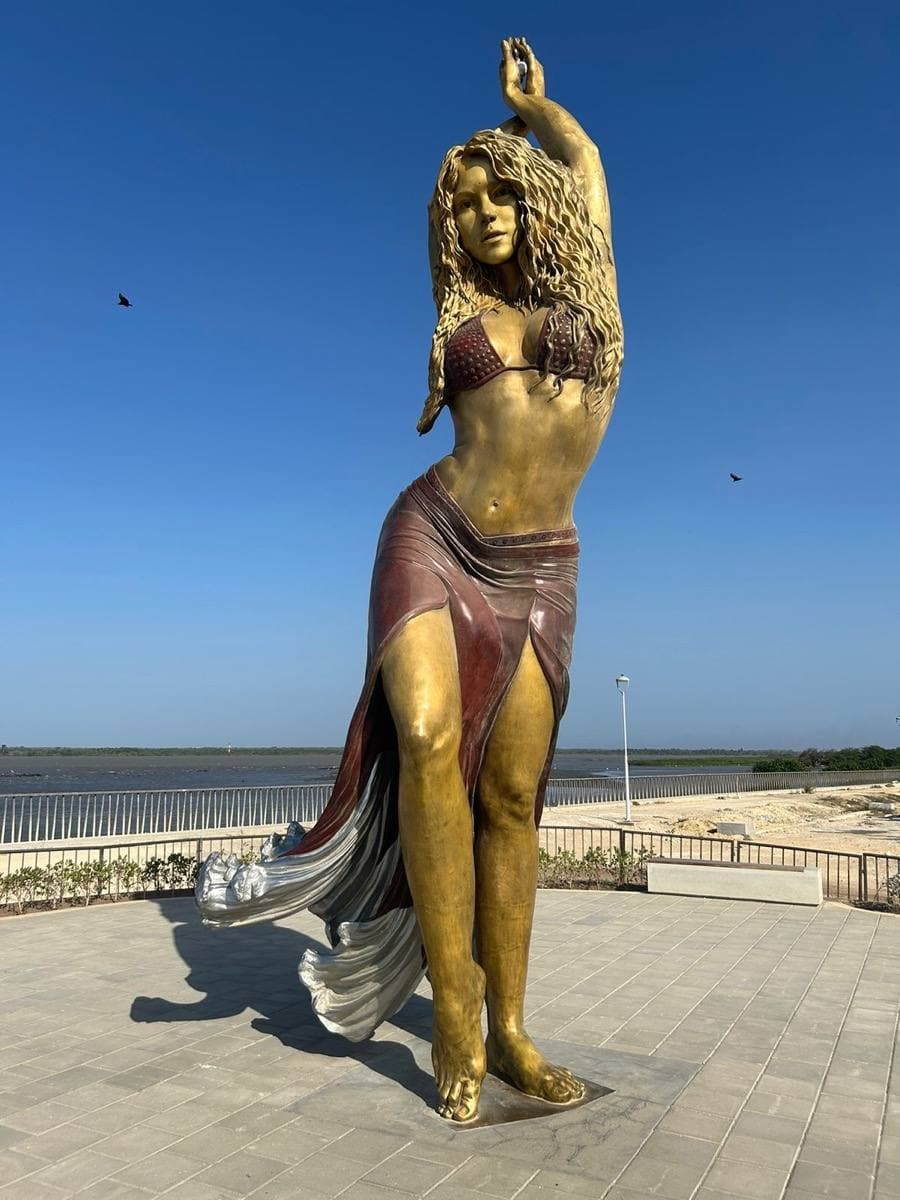 A tall bronze statue of Shakira with raised arms by the river
