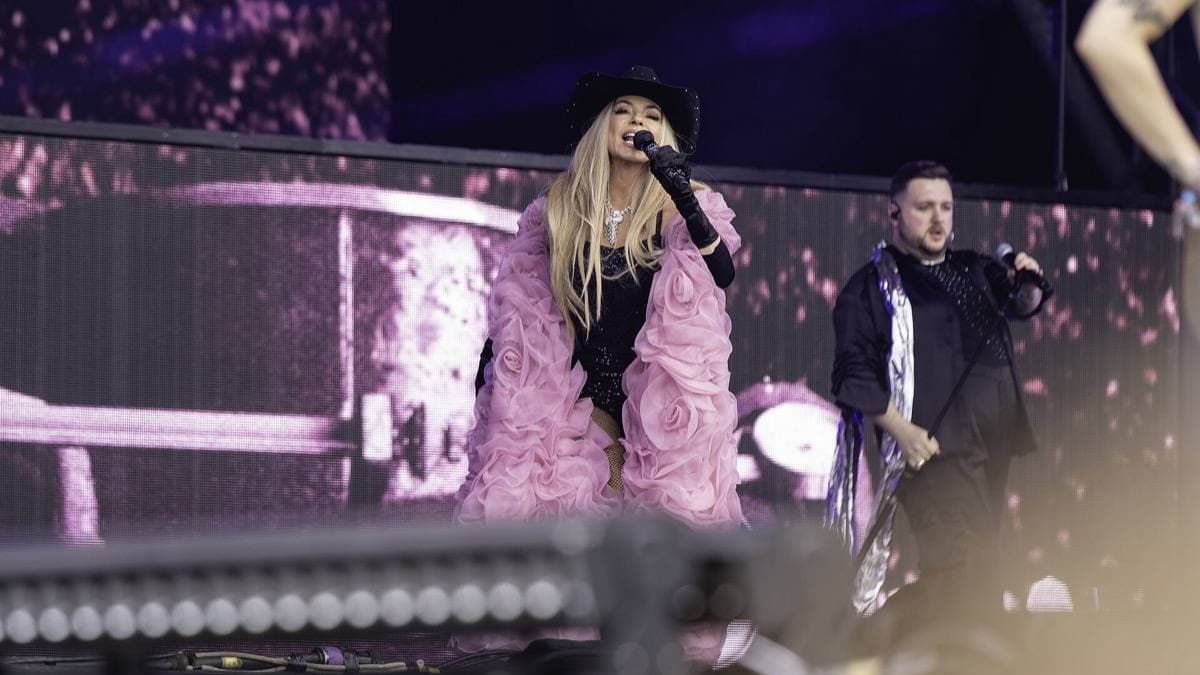 Shania Twain in a pink ruffled coat and black hat singing on a big festival stage