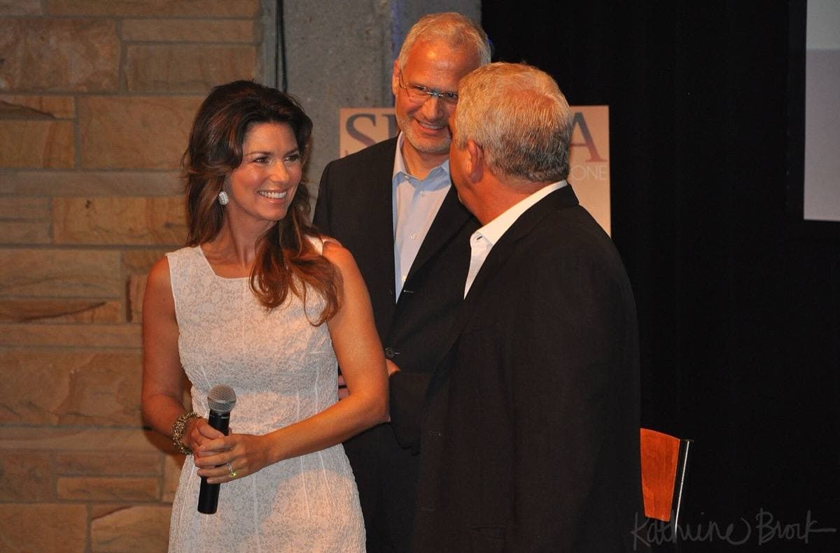 Shania Twain in a white dress holding a microphone beside two men at a press event