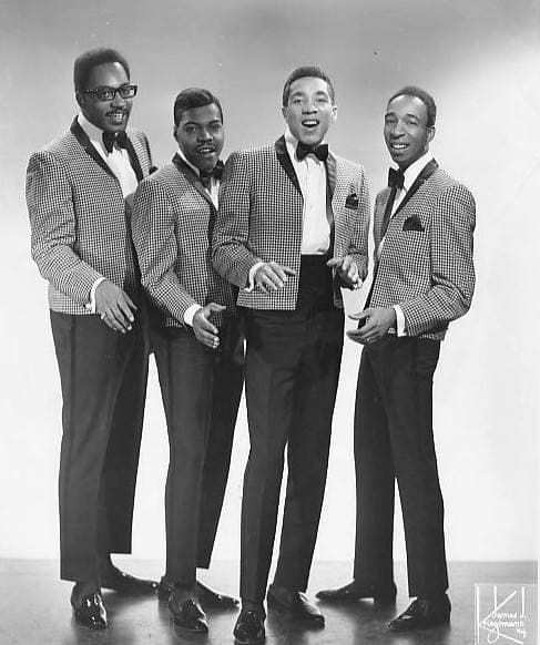 Four men in suits grouped closely, one in glasses, the front man smiling