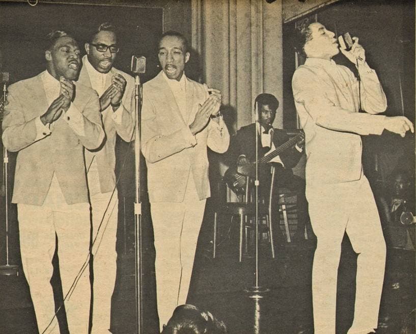 Three singers in pale suits clapping at microphones while the lead singer sings at the right