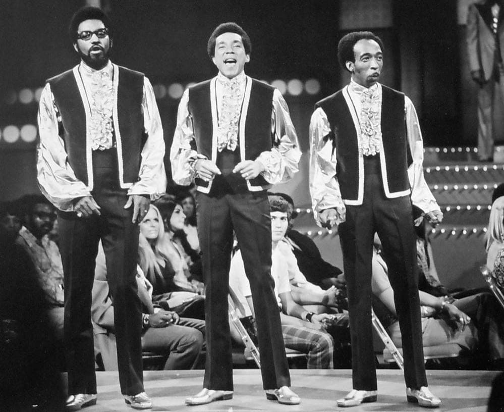 Three singers in waistcoats over ruffled satin shirts on a television stage with an audience behind