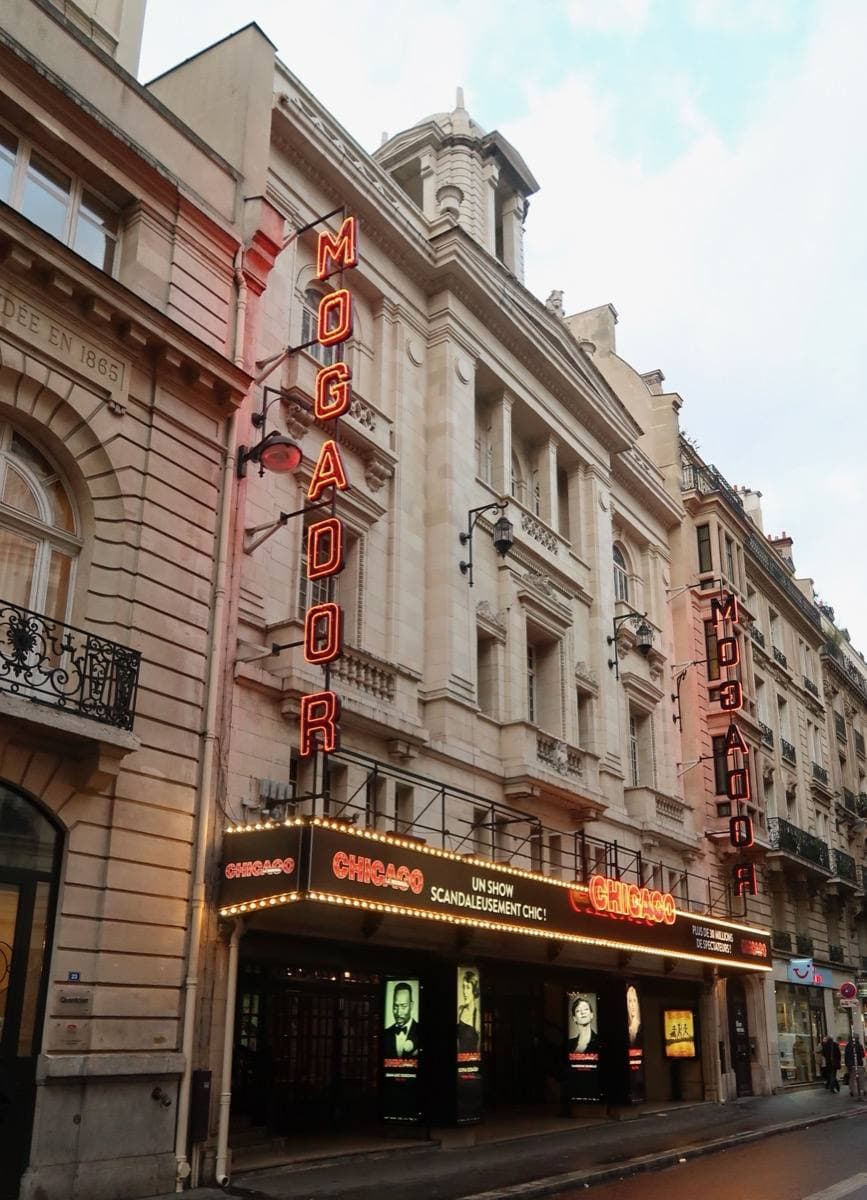 The tall stone front of a Paris theatre with a lit canopy