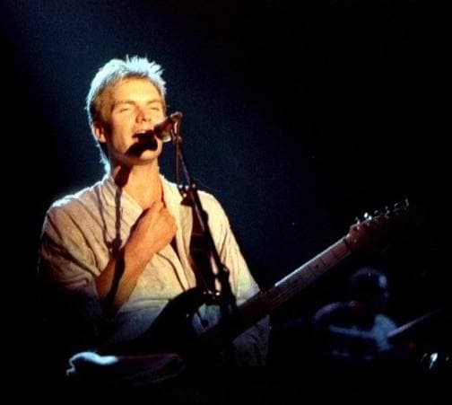 Sting on stage singing into a microphone, short fair hair, in stage light