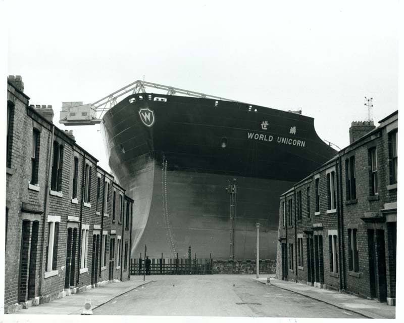 A tanker’s black bow towers over the end of a street of brick terraced houses, in black and white