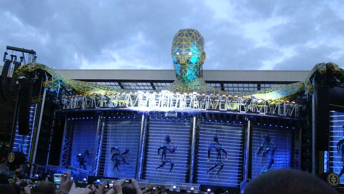 A huge faceted head rising over the top of a wide stadium stage under a grey sky