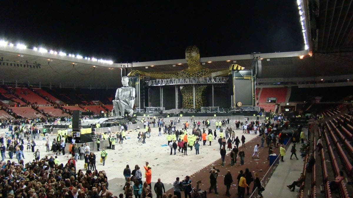 A seated giant figure on a stadium floor with the lights up and people heading for the exits