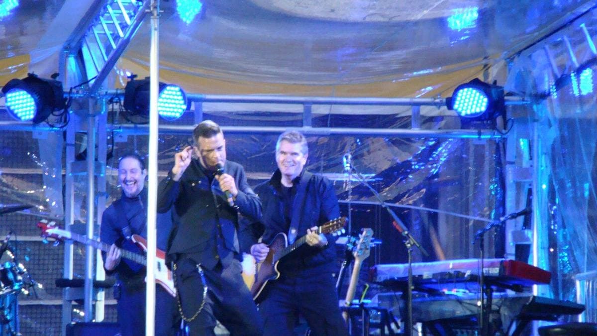 Robbie Williams singing in black with two guitarists under blue stage lights