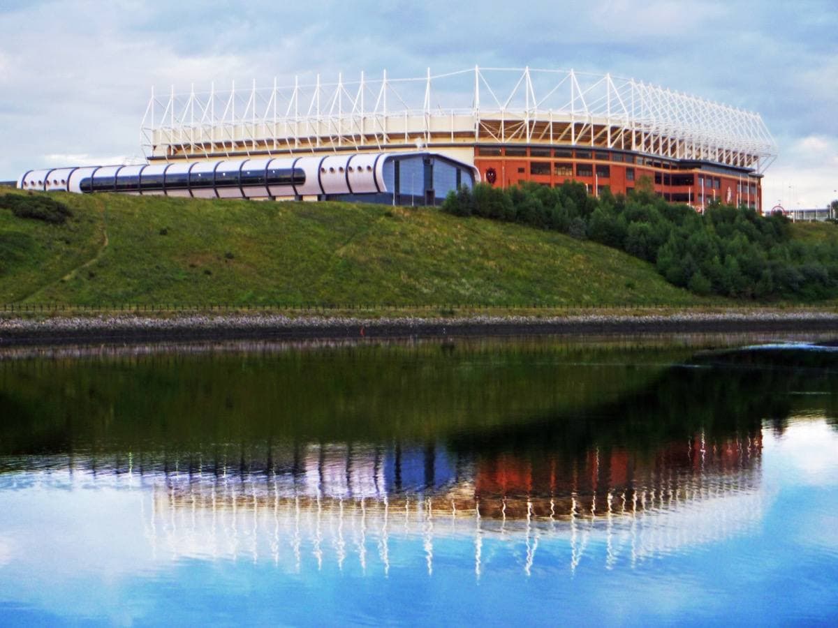 A red-brick stadium with a white roof reflected in still river water