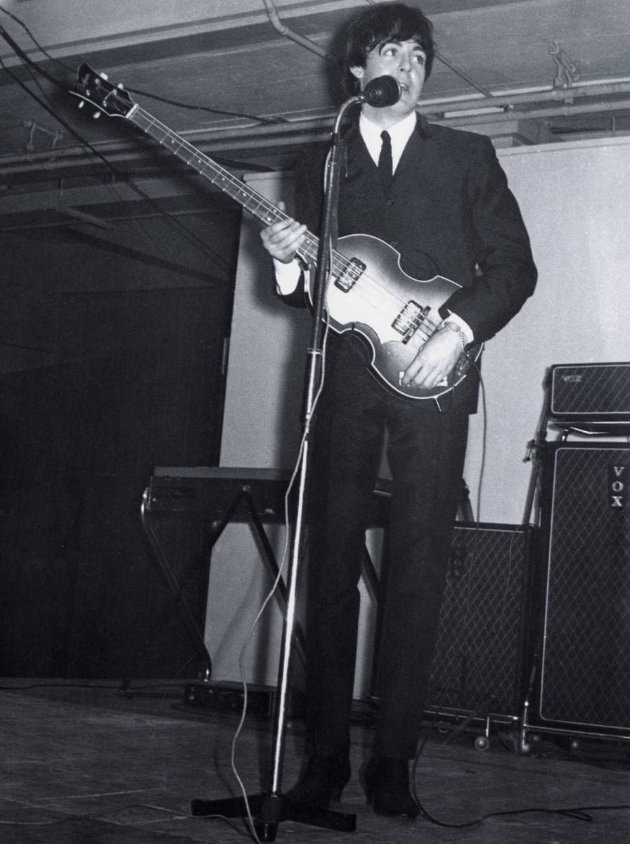 Paul McCartney in a dark suit playing a violin-shaped bass at a microphone