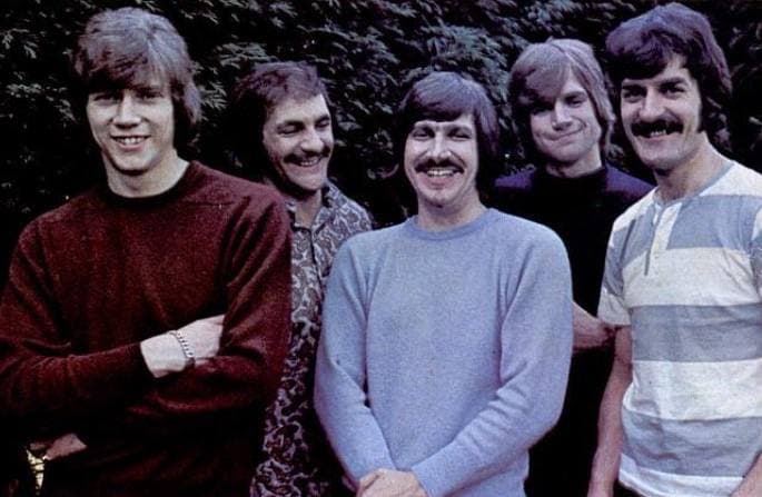The five band members smiling in jumpers in front of foliage, in colour, 1968