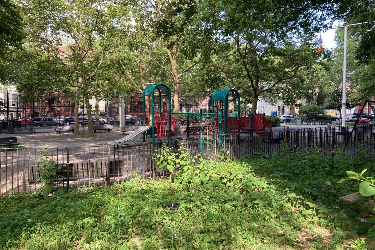 A fenced playground under tall trees with housing blocks behind