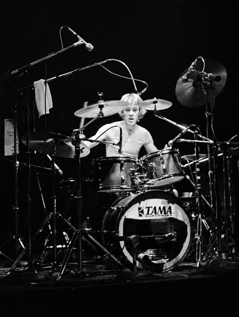 Stewart Copeland behind a large drum kit with many cymbals, in black and white