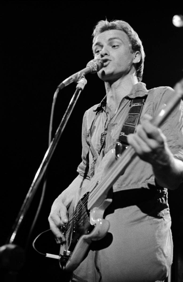 Sting singing with a bass guitar, short bleached hair, stage lights behind, in black and white