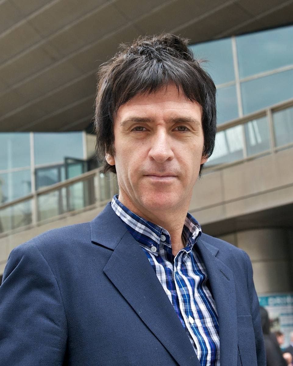 Johnny Marr in a navy jacket and checked shirt outside a glass building