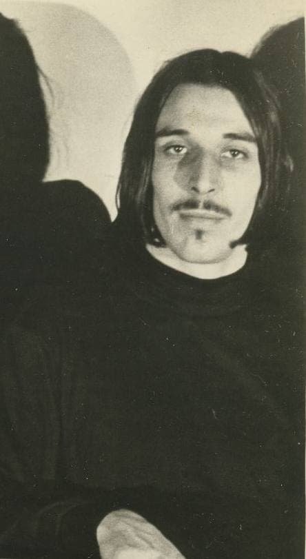 John Cale in black and white, long dark hair and a thin goatee, looking at the camera