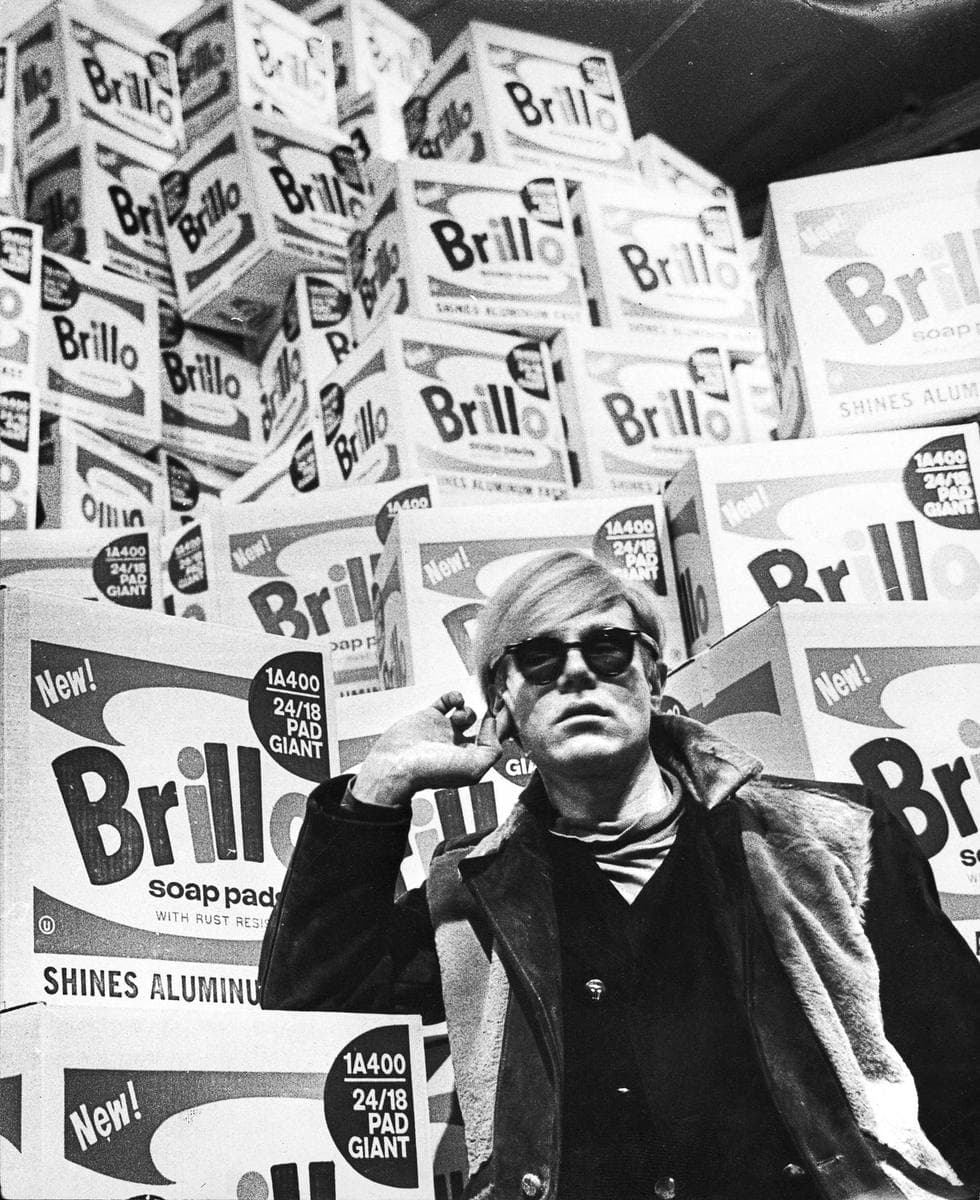 Andy Warhol in dark glasses and a leather jacket, Brillo boxes stacked behind him