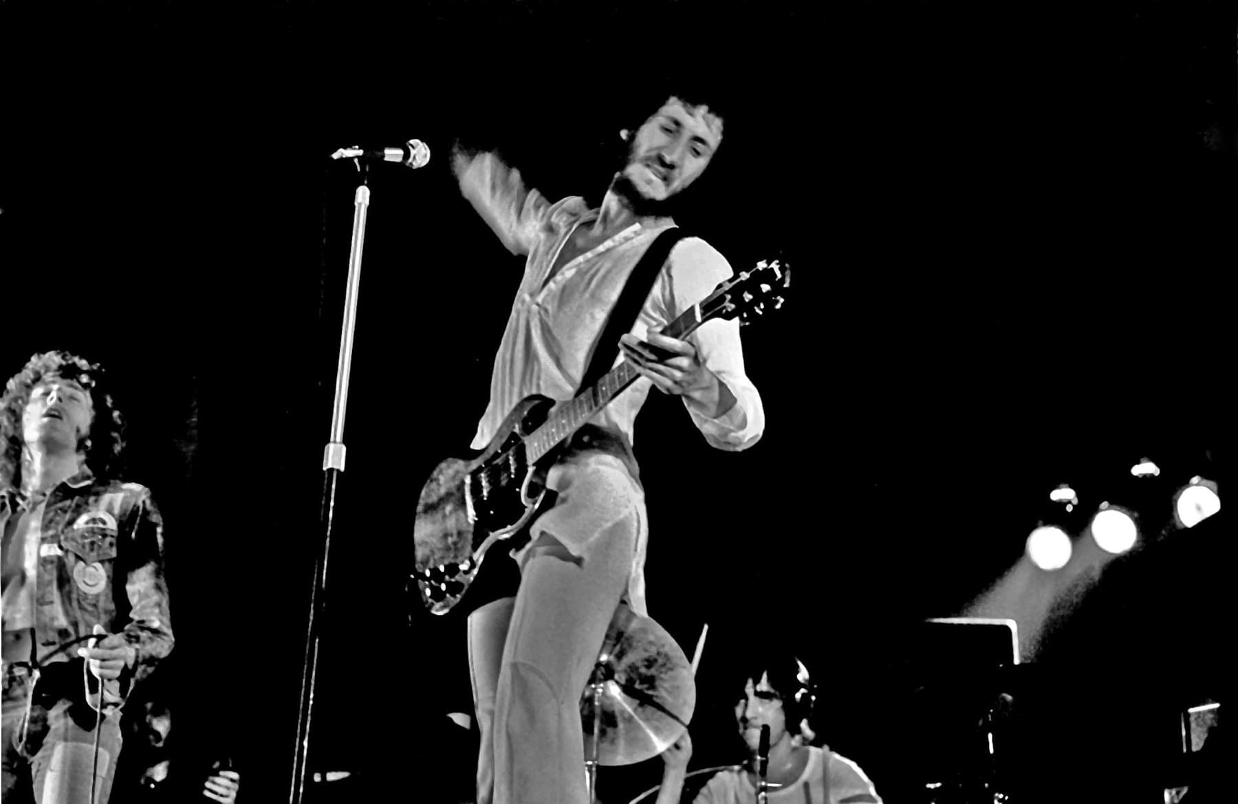 Pete Townshend mid-swing with a guitar under a stage light, Daltrey at the left and Moon at the drums