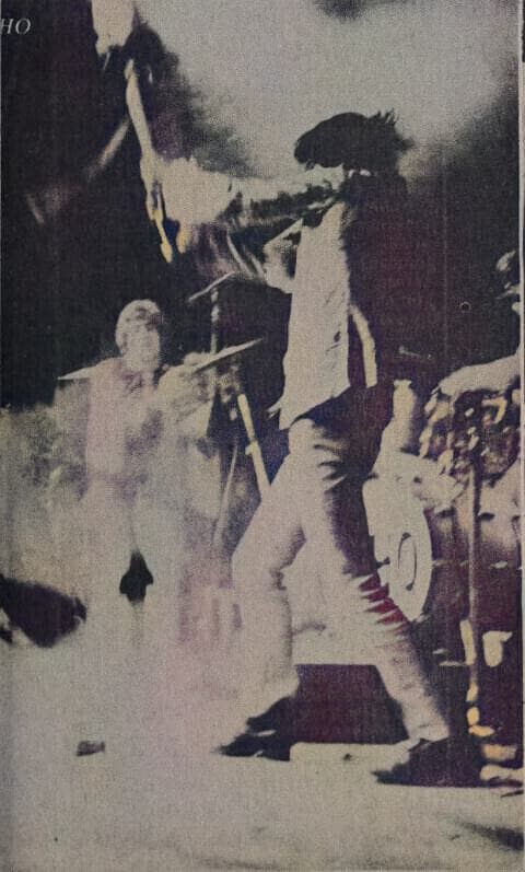 A grainy colour photograph of Pete Townshend swinging a guitar down on stage, Daltrey behind him