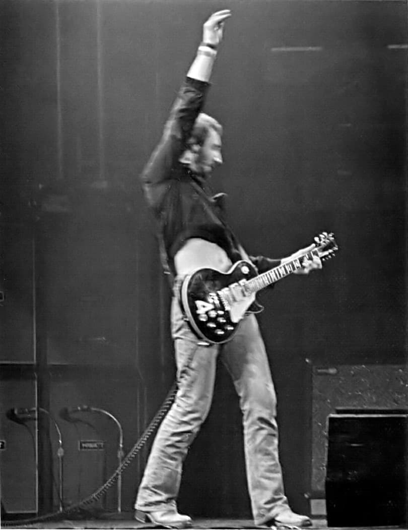 Pete Townshend on stage with his right arm straight up above a black guitar, amplifiers behind him