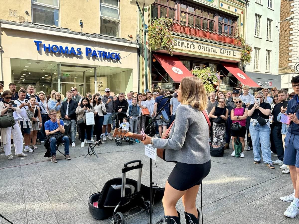 A crowd gathered round a guitarist on a pedestrian street below a café front with red awnings
