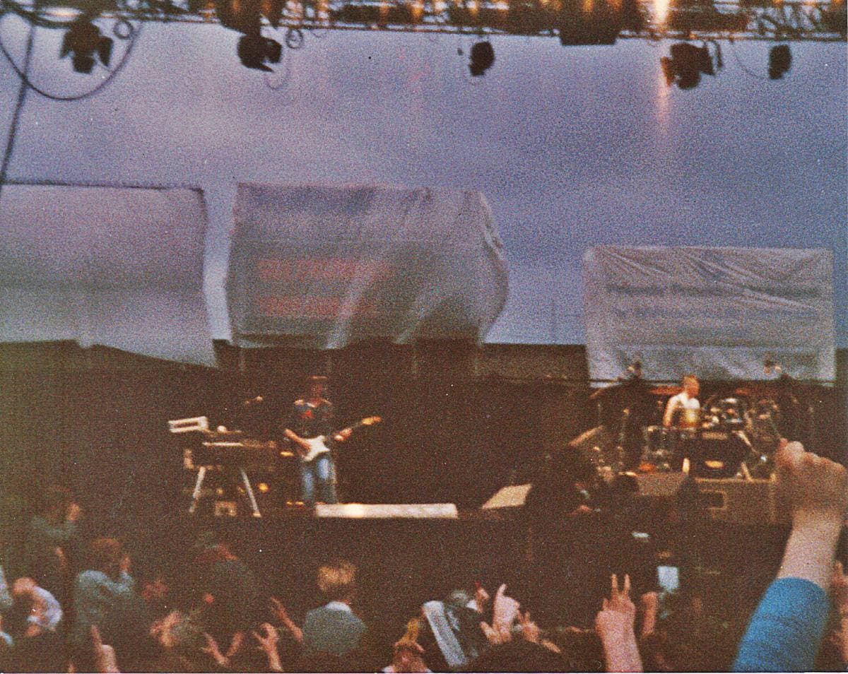 A stadium stage seen over a crowd with raised hands, banners hanging from the scaffold
