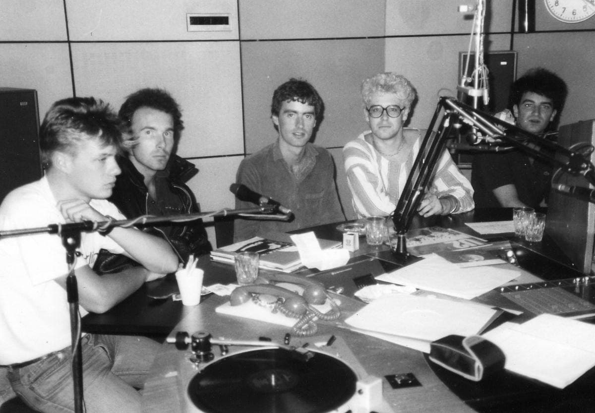 Five young men round a radio studio desk with microphones and a turntable, in black and white