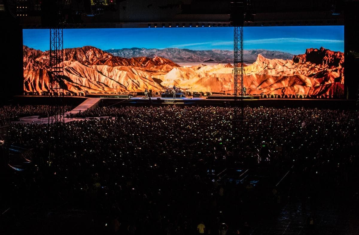 A stadium crowd in the dark below a vast screen showing orange desert badlands