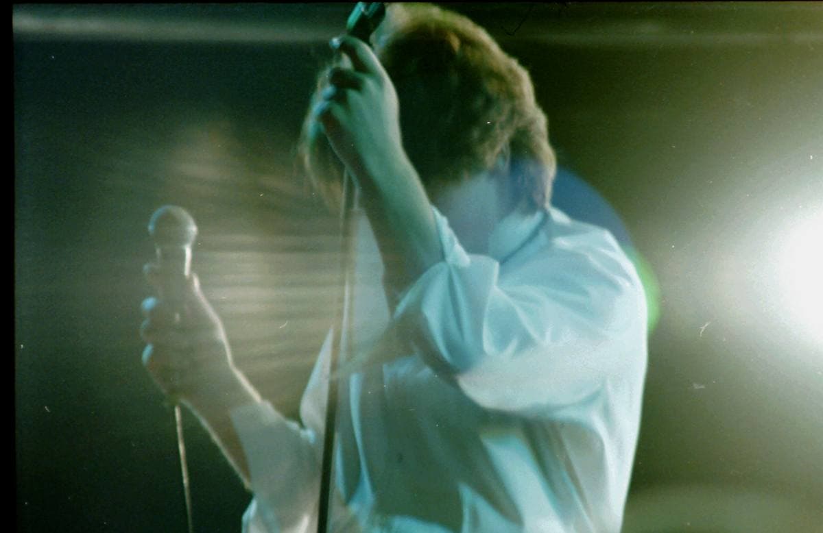 Bono in a white shirt holding the microphone stand, lit green and white