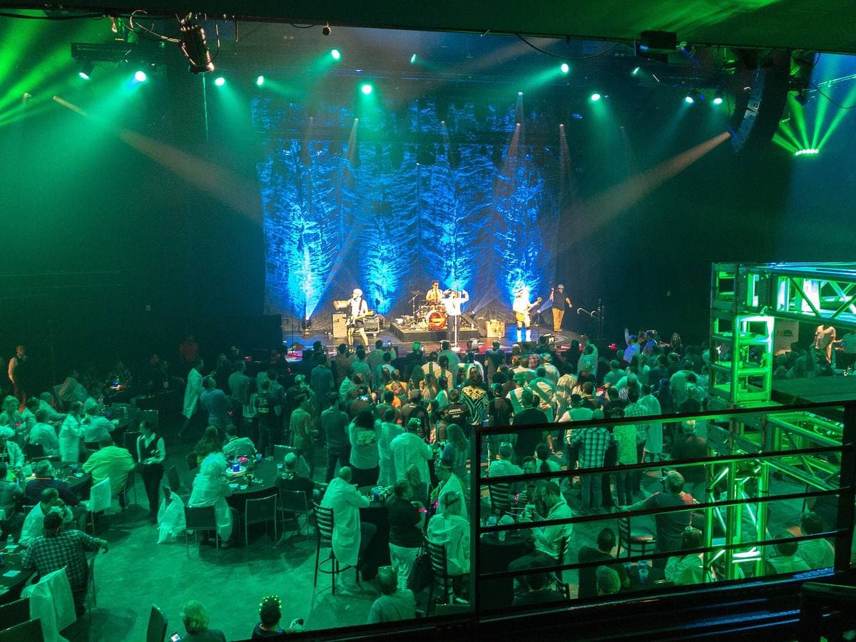 A packed theatre lit green and blue, a band on stage beyond the crowd