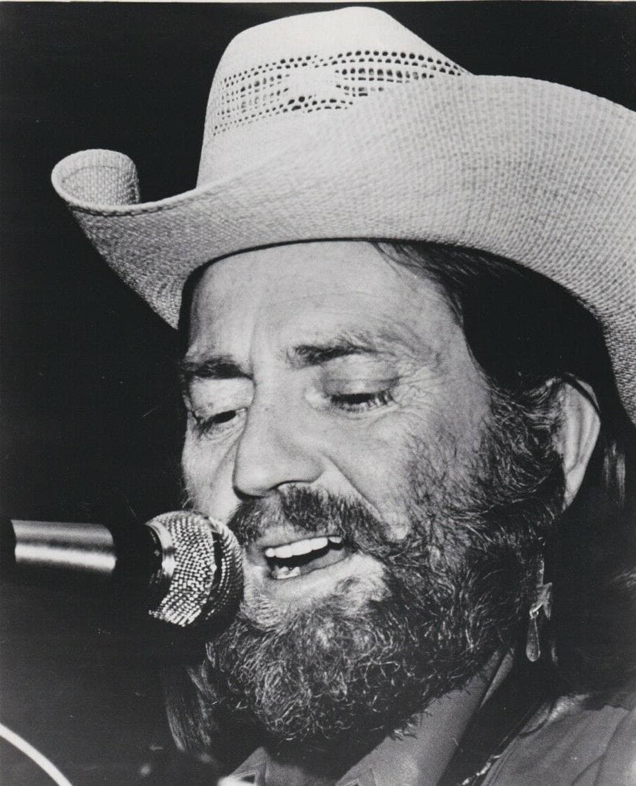 Willie Nelson in a straw cowboy hat, bearded, eyes half closed, singing at a microphone