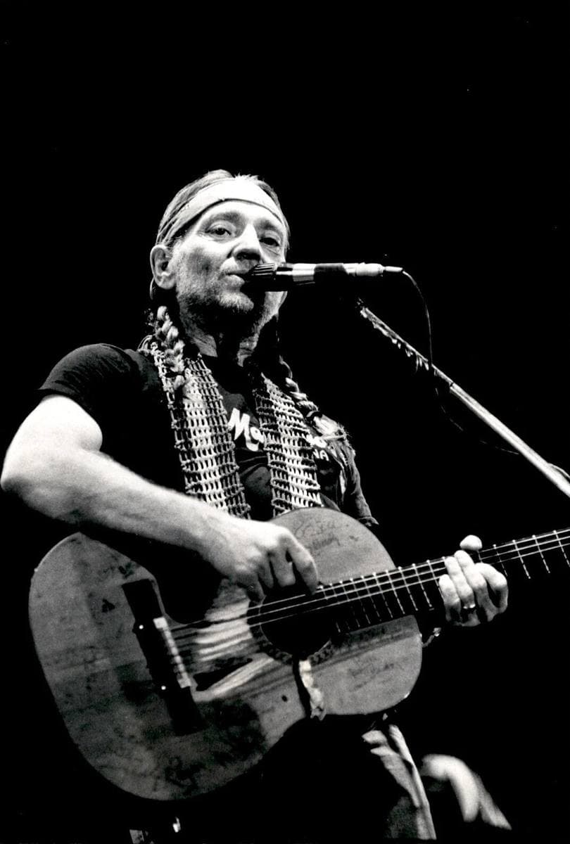 Willie Nelson singing into a microphone, a bandana round his head, playing a battered classical guitar covered in scratched names