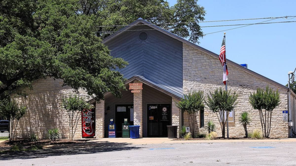 A stone and grey single-storey civic building with a flag and young trees in front