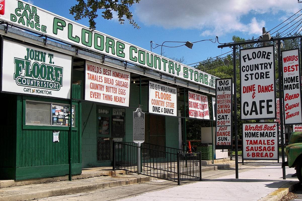 A green and white roadside building hung with hand-painted signs for a country store, cafe, dancing and tamales