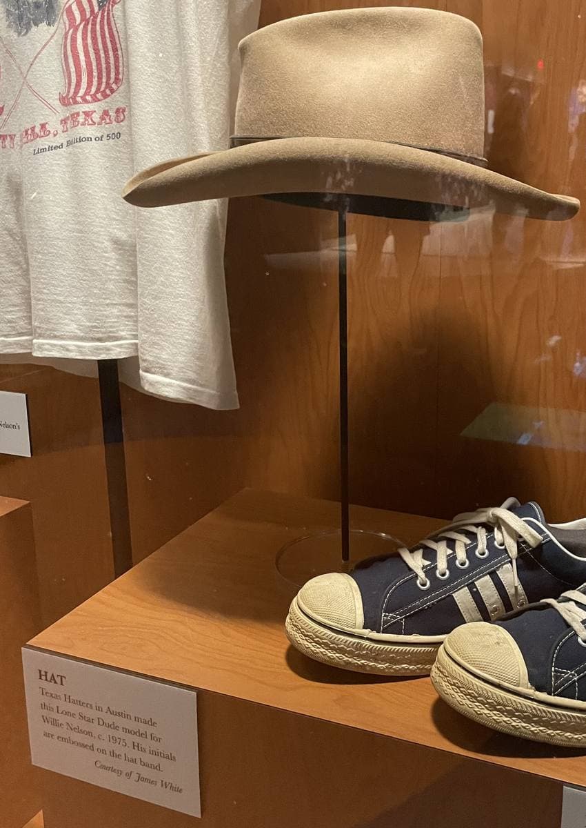 A pale cowboy hat on a stand in a museum case above a pair of canvas sneakers and a placard