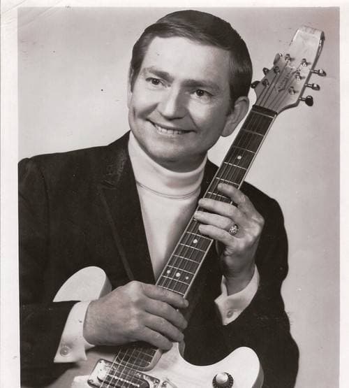 Willie Nelson in a dark jacket and white roll-neck, smiling, holding an electric guitar