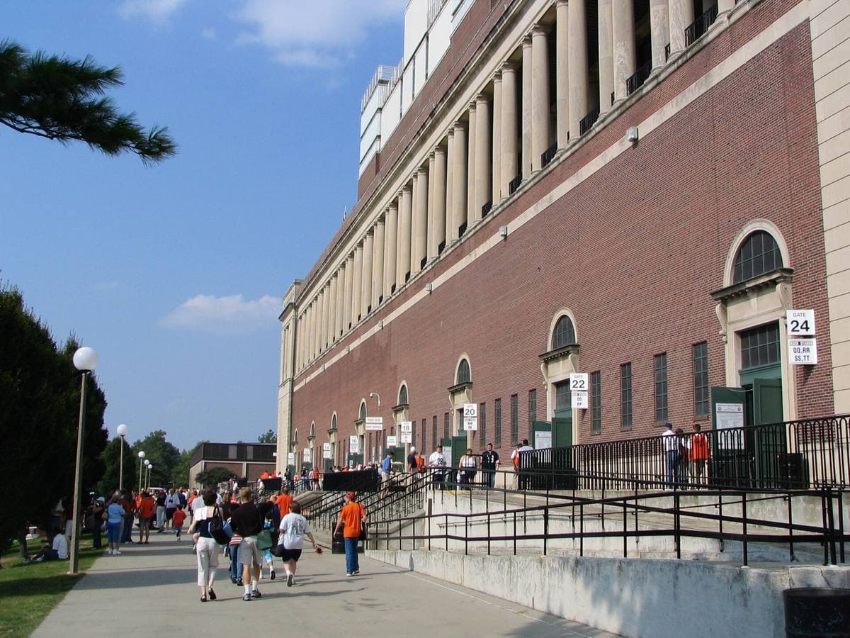 The long brick and stone colonnaded front of a football stadium with fans walking past
