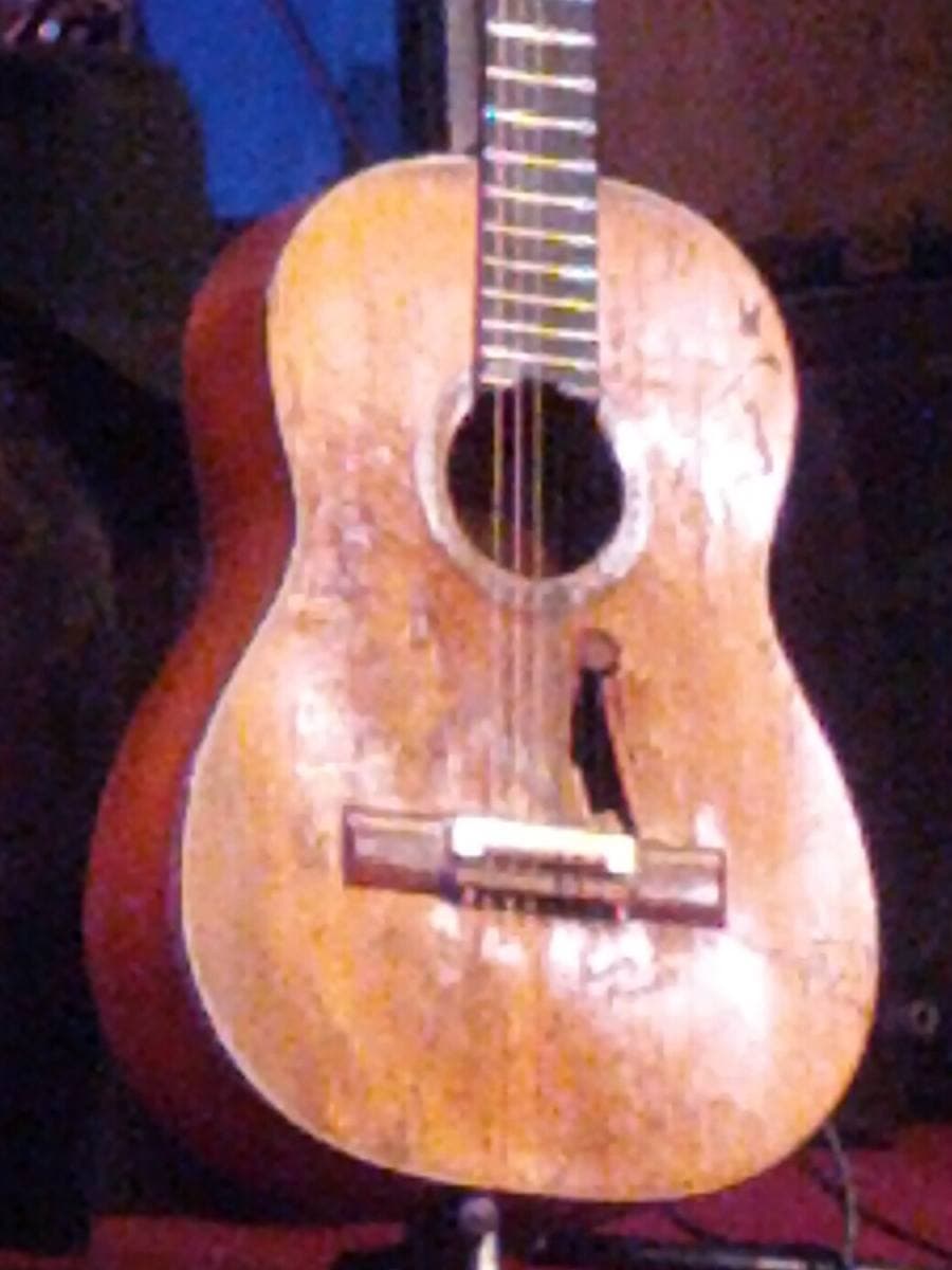 A battered classical guitar on a stand on a stage, its top worn pale with a hole under the sound hole