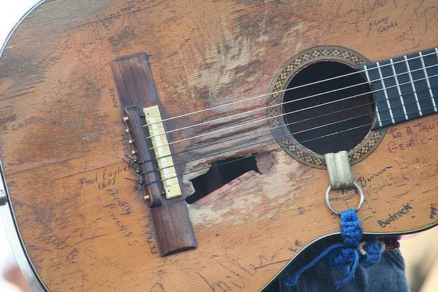 Close-up of a worn nylon-string guitar top with a ragged hole below the strings and handwritten names scratched across the wood