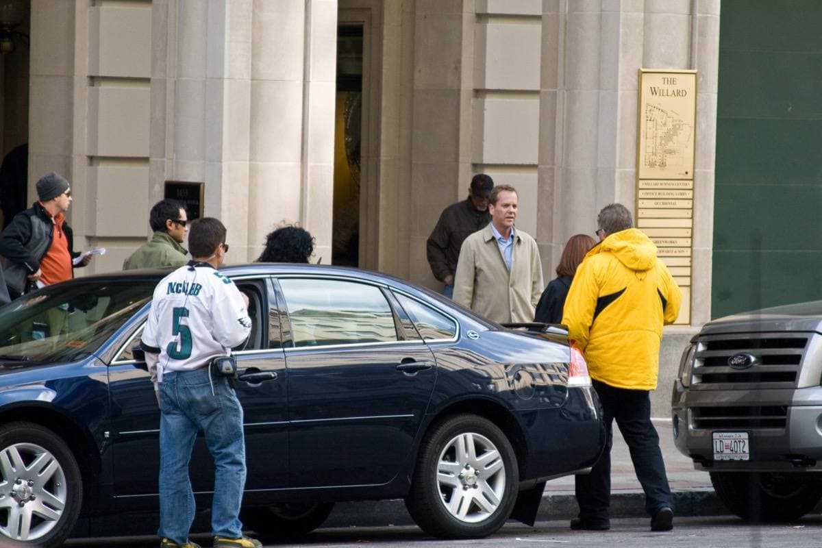 Kiefer Sutherland in a dark jacket on a city street during filming, crew and equipment around him
