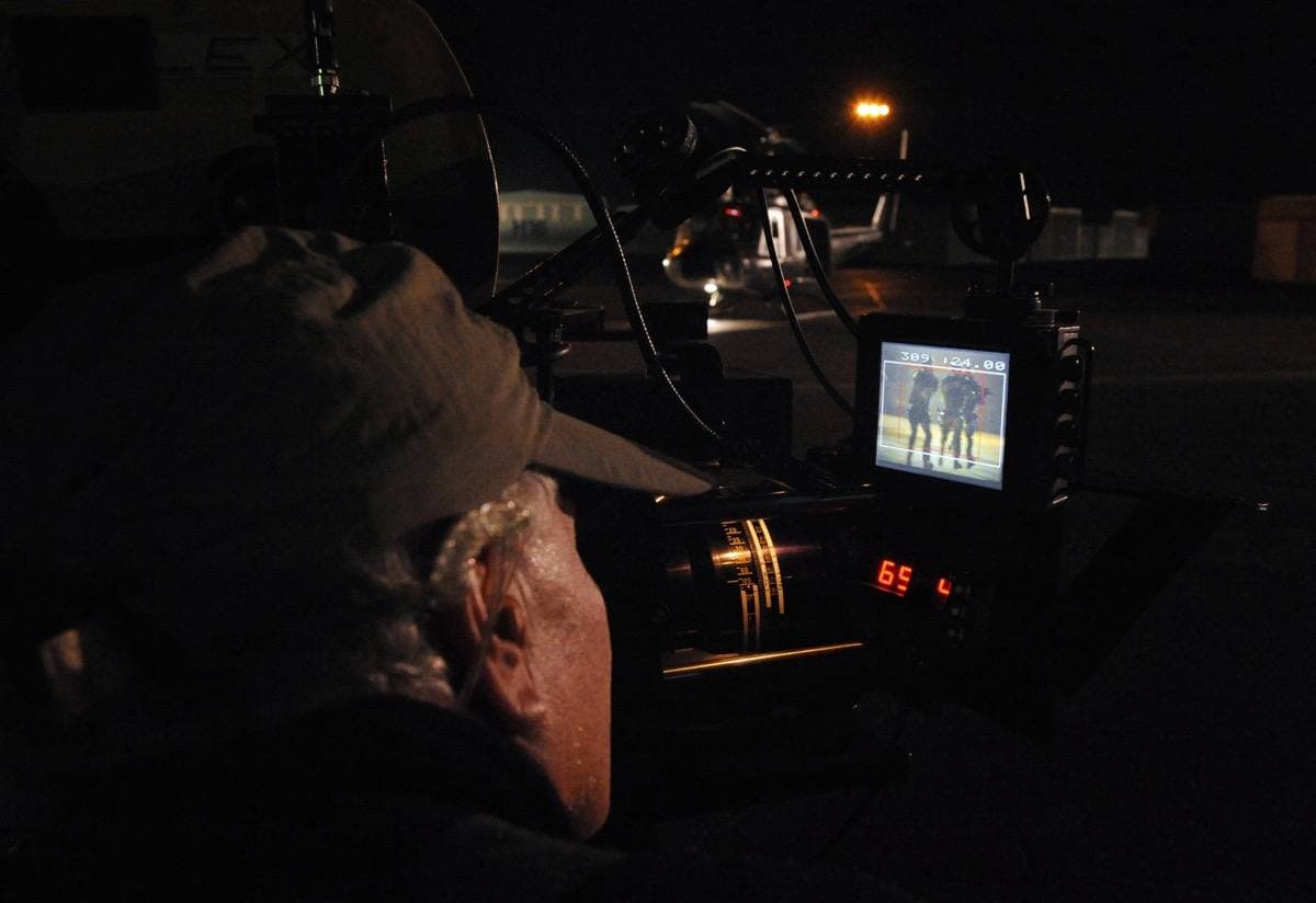 A camera operator filming men in uniform beside a helicopter on an airfield