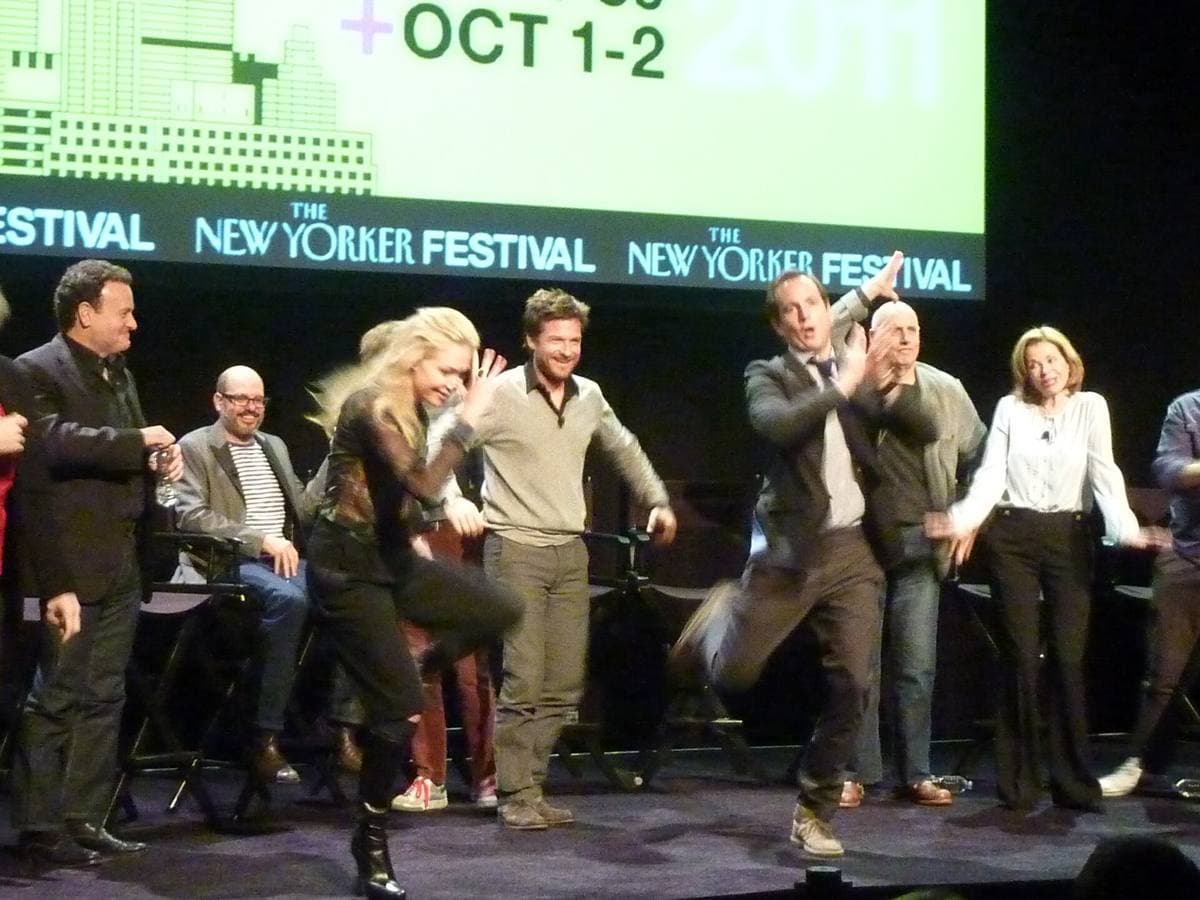 The cast of Arrested Development on a stage under a New Yorker Festival screen, flapping their arms like chickens