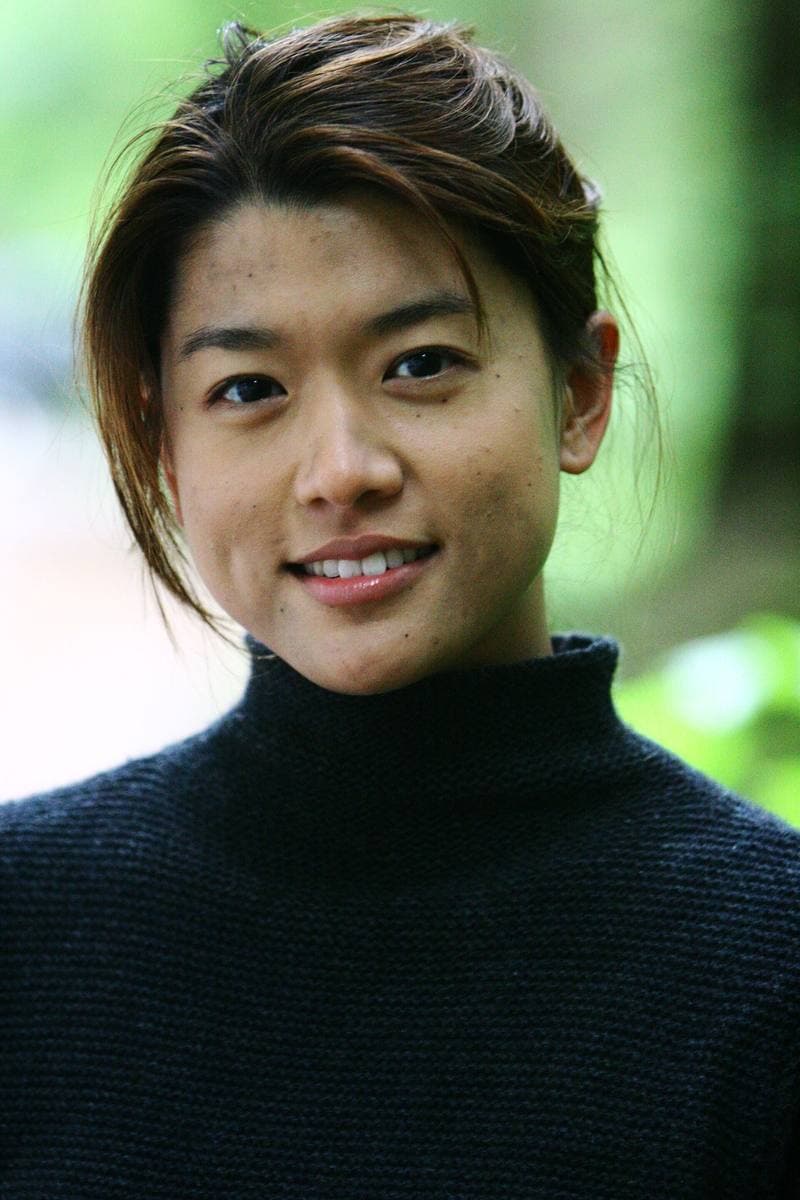 A young woman in a dark knitted jumper, smiling, with forest behind her